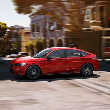 2026 Honda Civic Hatchback Sport Hybrid in Rallye Red profile driving on a city street (Compact Hatchback).