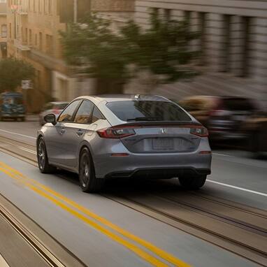2026 Honda Civic Hatchback Sport Hybrid in Urban Gray Pearl back driving on a city street (Compact Hatchback).
