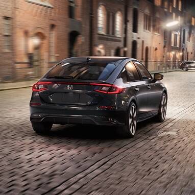 2026 Honda Civic Hatchback Sport Touring Hybrid in Meteorite Gray Metallic back driving on a cobblestone city street (Compact Hatchback).